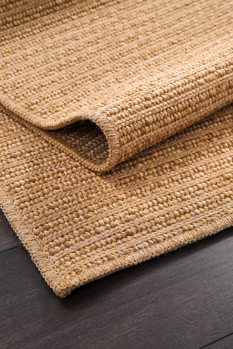 May include: A natural fiber rug with a rolled edge. The rug is made of woven, light brown fibers, with a textured surface and a border. The rug is partially rolled up, revealing the underside and the woven construction.