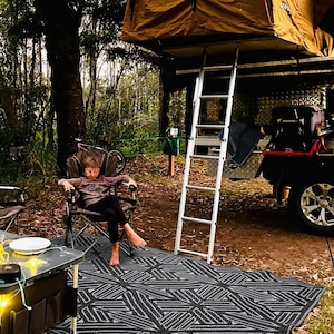 Pode incluir: Uma crian&ccedil;a est&aacute; sentada em uma cadeira de acampamento em um tapete de exterior estampado em preto e branco. Uma escada leva a uma barraca de lona marrom no topo de um ve&iacute;culo.