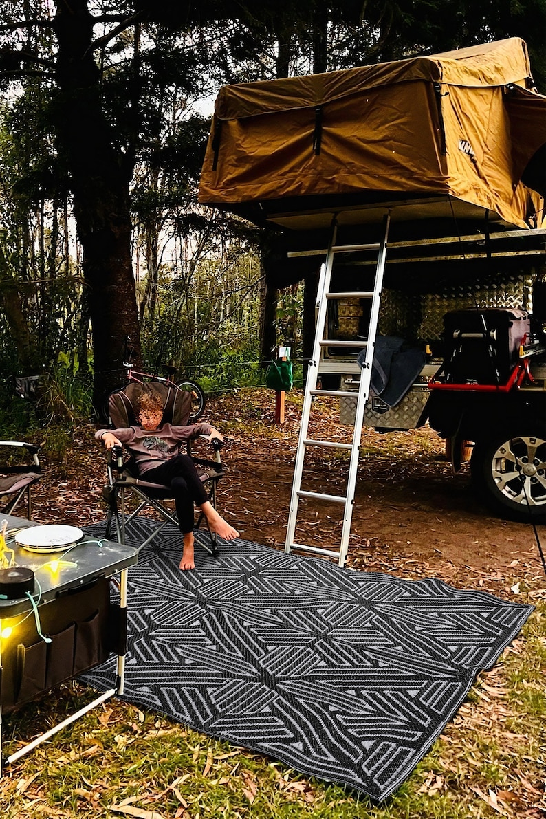 Pode incluir: Uma crian&ccedil;a est&aacute; sentada em uma cadeira de acampamento em um tapete de exterior estampado em preto e branco. Uma escada leva a uma barraca de lona marrom no topo de um ve&iacute;culo.