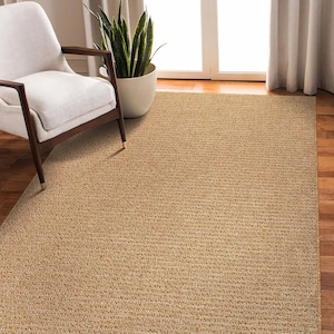 May include: A rectangular, woven area rug in a natural tan color. The rug has a textured surface and is placed on a hardwood floor. A white chair with dark wooden legs and a potted plant are in the background.