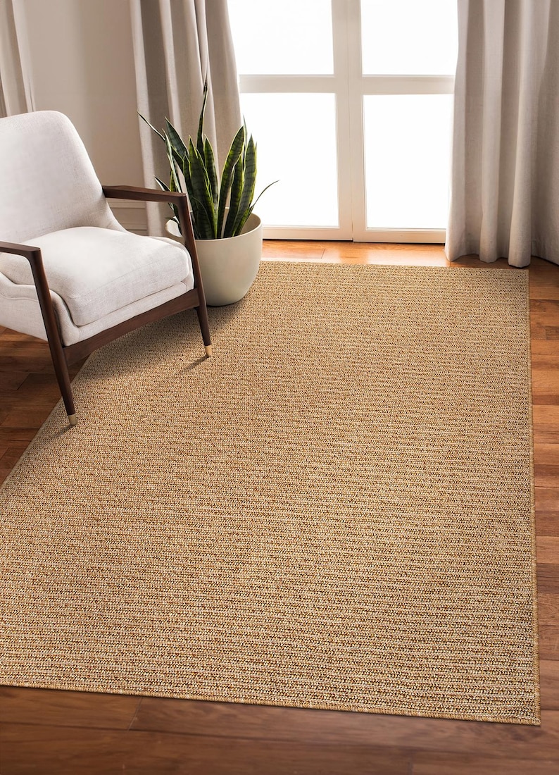 May include: A rectangular, woven area rug in a natural tan color. The rug has a textured surface and is placed on a hardwood floor. A white chair with dark wooden legs and a potted plant are in the background.