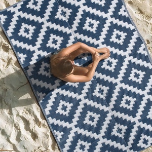 May include: A blue and white patterned outdoor rug with a geometric design. The rug is laid out on a sandy beach.