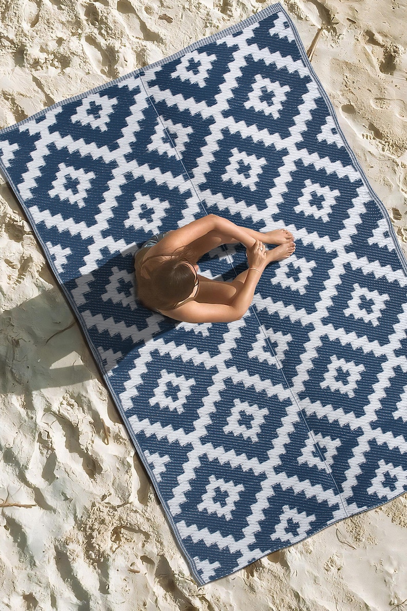 May include: A blue and white patterned outdoor rug with a geometric design. The rug is laid out on a sandy beach.