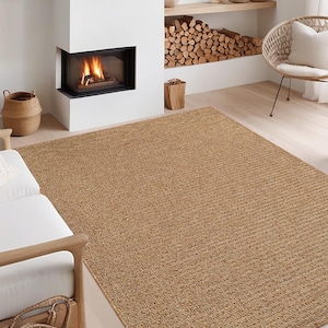 May include: A large, rectangular, woven jute rug in a warm tan color. The rug has a textured surface with a linear pattern. The rug is placed in a living room setting with a fireplace and wicker chair.