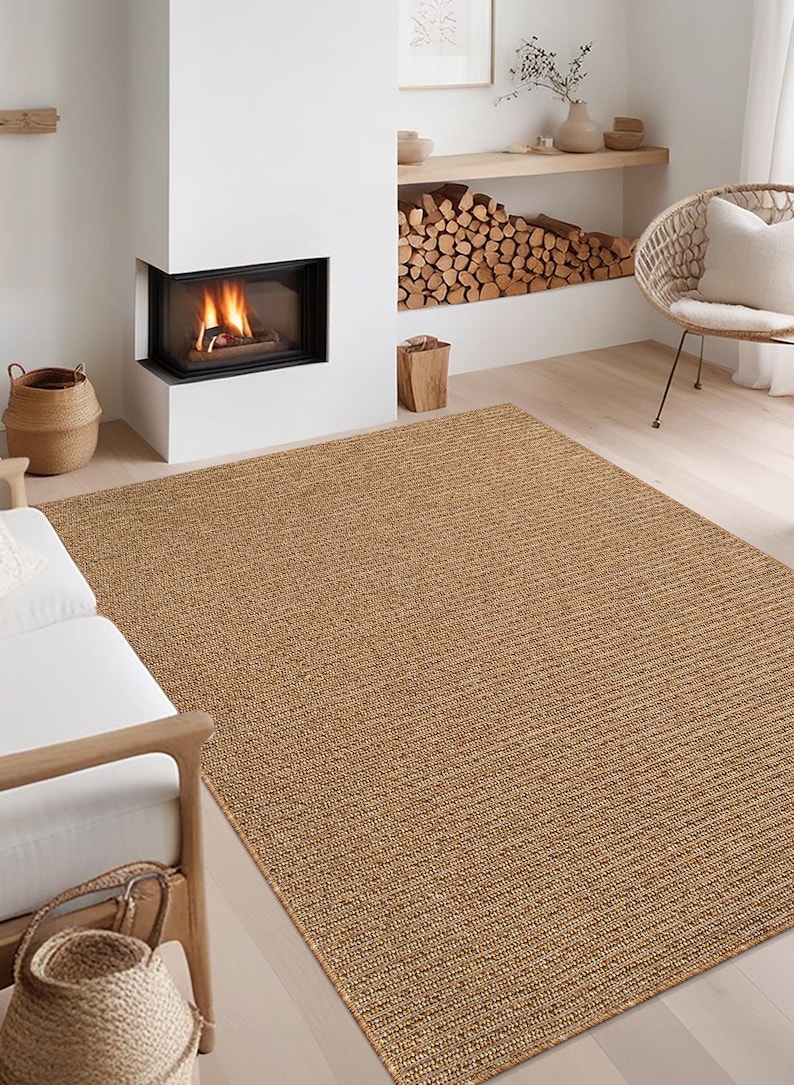 May include: A large, rectangular, woven jute rug in a warm tan color. The rug has a textured surface with a linear pattern. The rug is placed in a living room setting with a fireplace and wicker chair.