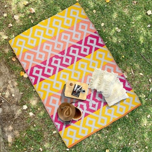 Könnte beinhalten: Eine geometrisch gemusterte Picknickdecke in Gelb, Rosa und Grau mit einem Buch, zwei Tassen, einem Hut und zwei weißen Kissen darauf.