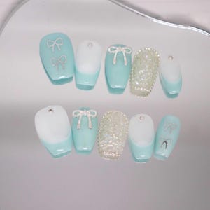 May include: A set of ten light blue coffin-shaped press-on nails with different designs. Some nails have a white tip, some have silver bows, and some have a sparkly design.