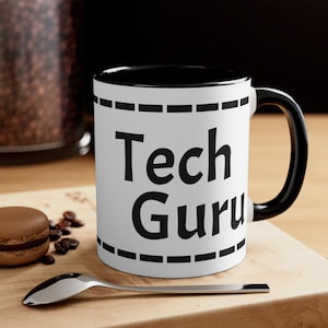 Puede incluir: Taza de cerámica blanca con borde y asa negros. La taza tiene texto negro que dice "Tech Guru" con una línea discontinua negra arriba y abajo del texto.