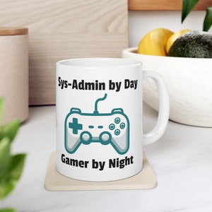 Könnte beinhalten: Weiße Keramiktasse mit einem schwarzen und türkisfarbenen Gamepad-Motiv und dem Text "Sys-Admin by Day Gamer by Night".