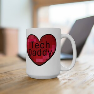 Pode incluir: Caneca de cerâmica branca com um grande design de coração vermelho. O coração tem um padrão pixelizado e as palavras "Tech Daddy" em preto. A caneca tem uma alça grande e está sobre uma superfície de madeira.
