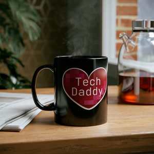 May include: Black coffee mug with a red heart design that says "Tech Daddy" in white letters. The mug is sitting on a wooden table with a stack of newspapers.