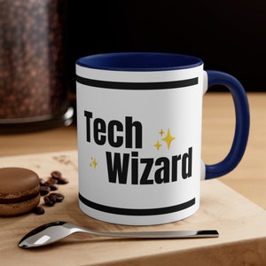 May include: White ceramic mug with a blue rim and handle. The mug has black text that reads "Tech Wizard" with gold stars on either side of the text.