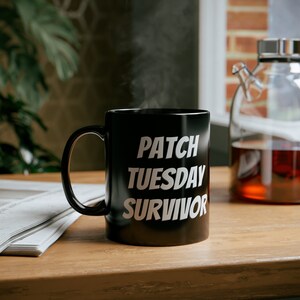 May include: A black ceramic mug with white text that reads "PATCH TUESDAY SURVIVOR". The mug is filled with a hot drink and steam is rising from the top.