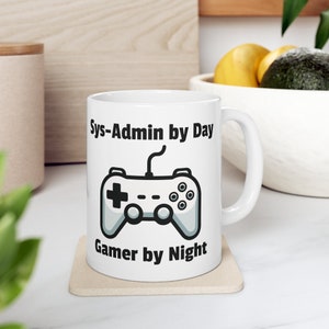 Könnte beinhalten: Weiße Keramiktasse mit einem schwarzen und grauen Videospiel-Controller-Motiv. Der Text "Sys-Admin by Day Gamer by Night" ist auf die Tasse gedruckt.