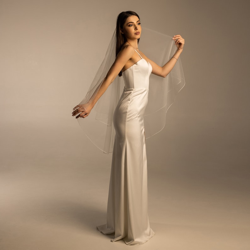 May include: A woman wearing a white satin wedding dress with a long, single-tiered veil. The veil is draped over her head and shoulders, and she is holding the edge of the veil in her right hand.