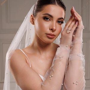 K&ouml;nnte beinhalten: Eine Braut mit einem wei&szlig;en Schleier und durchsichtigen Handschuhen, die mit Perlen verziert sind. Der Schleier ist &uuml;ber Kopf und Schultern drapiert, die Handschuhe reichen bis zu den Ellbogen. Das Bild ist eine Nahaufnahme.