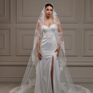 May include: A white satin wedding dress featuring a sweetheart neckline and a high slit. The dress is complemented by a long, sheer veil edged with intricate lace. The veil trails to the floor, adding a touch of elegance.