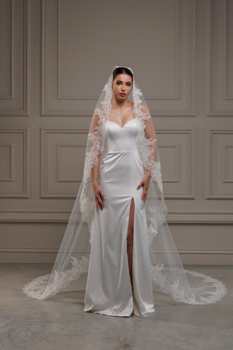 May include: A white satin wedding dress featuring a sweetheart neckline and a high slit. The dress is complemented by a long, sheer veil edged with intricate lace. The veil trails to the floor, adding a touch of elegance.