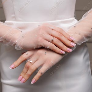 K&ouml;nnte beinhalten: Nahaufnahme eines wei&szlig;en Brautkleides mit durchsichtigen, langen Handschuhen. Die Handschuhe sind mit kleinen, glitzernden Verzierungen versehen. Die N&auml;gel sind mit einer rosa-wei&szlig;en French Manik&uuml;re lackiert. Das Bild hat den Text "LIKE VEIL".