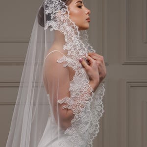 May include: A white bridal veil with detailed floral lace edging. The sheer veil drapes over the shoulders, creating an elegant and romantic appearance. The bride is wearing a white dress.