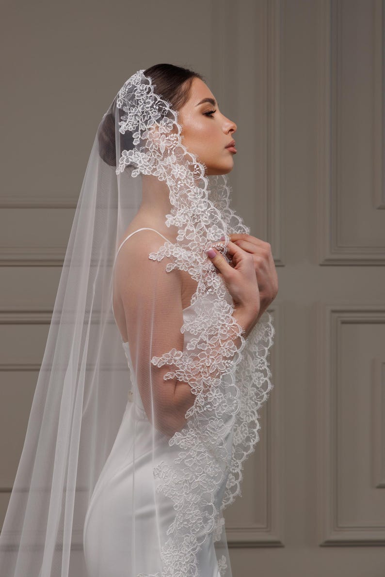 May include: A white bridal veil with detailed floral lace edging. The sheer veil drapes over the shoulders, creating an elegant and romantic appearance. The bride is wearing a white dress.