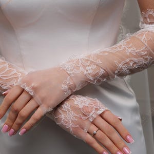K&ouml;nnte beinhalten: Elegante wei&szlig;e Brauthandschuhe aus Spitze. Die Handschuhe weisen aufwendige florale Spitzendesigns auf und reichen bis zum mittleren Unterarm. Das zarte Design verleiht Hochzeitskleidung einen Hauch von Raffinesse. Die Handschuhe sind aus einem transparenten, leichten Stoff gefertigt.