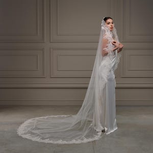 May include: A white bridal veil with a detailed lace edge and a long train. The veil is worn with a white satin wedding dress. The bride is standing in front of a neutral wall.