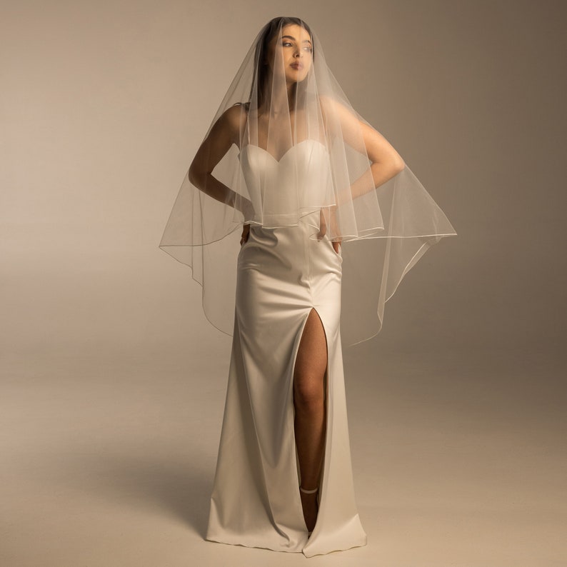 May include: A woman wearing a white wedding dress with a long veil. The dress has a thigh-high slit and a fitted bodice.
