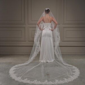 May include: A bride in a white wedding dress and veil, seen from behind. The dress features a fitted bodice and a flowing skirt. The veil is long, with a delicate lace trim. The background is a neutral wall.