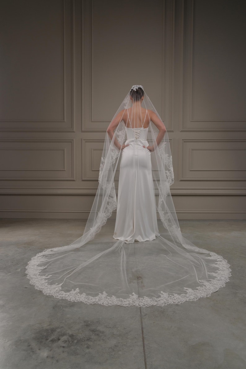 May include: A bride in a white wedding dress and veil, seen from behind. The dress features a fitted bodice and a flowing skirt. The veil is long, with a delicate lace trim. The background is a neutral wall.