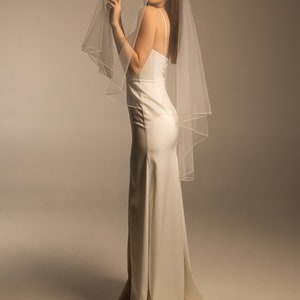 May include: A woman wearing a white wedding dress with a long veil. The veil is sheer and has a satin trim.