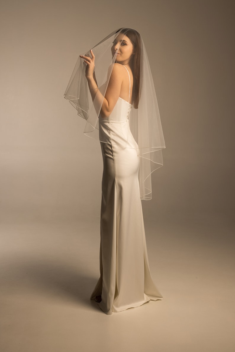 May include: A woman wearing a white wedding dress with a long veil. The veil is sheer and has a satin trim.
