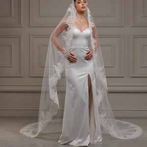 May include: A white satin wedding dress with a sweetheart neckline and a high slit. The dress is paired with a long, sheer veil trimmed with delicate lace. The bride is posing with her hands on her hips.