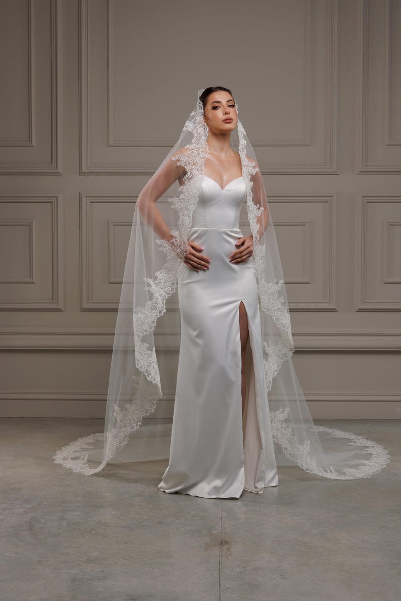 May include: A white satin wedding dress with a sweetheart neckline and a high slit. The dress is paired with a long, sheer veil trimmed with delicate lace. The bride is posing with her hands on her hips.