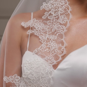 May include: A white bridal veil with delicate floral lace trim. The lace detail cascades down the shoulder and onto a white satin wedding dress. The veil is sheer, and the lace is intricately designed.