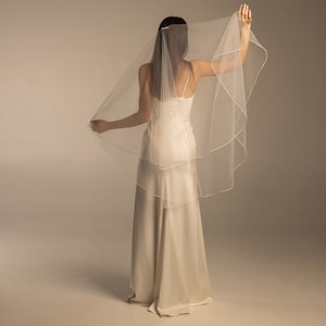 May include: A white bridal veil with a single layer of tulle and a satin edge. The veil is draped over the back of the bride's head and falls down to her shoulders.