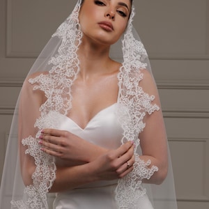 May include: A white bridal veil with intricate floral lace edging. The veil is draped over a white satin wedding dress. The bride is wearing makeup and a ring. The overall theme is bridal wear.