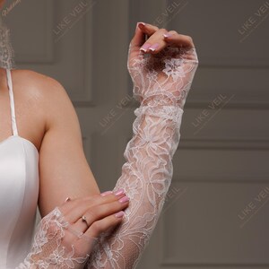 K&ouml;nnte beinhalten: Wei&szlig;e Brauthandschuhe aus Spitze mit floralen Mustern. Die Handschuhe reichen bis zum Oberarm und sind aus einem zarten, transparenten Stoff gefertigt. Das Bild zeigt auch ein wei&szlig;es Brautkleid und den Text "LIKE VEIL".