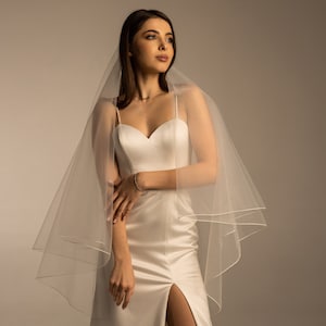 May include: A white bridal veil with a single layer of tulle and a satin trim. The veil is draped over the shoulders of a person wearing a white satin dress.