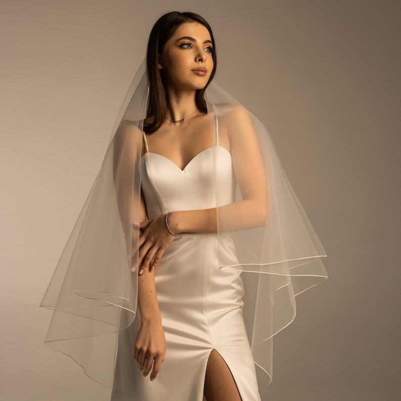 May include: A white bridal veil with a single layer of tulle and a satin trim. The veil is draped over the shoulders of a person wearing a white satin dress.