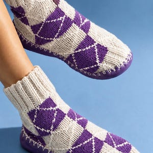 Könnte beinhalten: Ein Paar knöchelhohe Stricksocken mit einem violett-cremefarbenen Rautenmuster und weißen Ziernähten. Die Socken sind mit einer durchgehenden violetten Sohle verbunden, die am Rand sichtbare weiße Nähte aufweist, ähnlich einem Hausschuh.