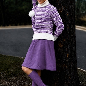 Könnte beinhalten: Ein violett-weiß gemusterter Rollkragenpullover mit passender langer Zipfelmütze und weißem Bommel, kombiniert mit einem einfarbigen violetten Rock, violetten Kniestrümpfen und schwarzen Schuhen. Der Pullover hat Querstreifen und weiße Bündchen.