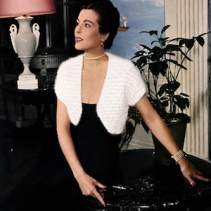 May include: A white fuzzy short-sleeved bolero cardigan worn over a black dress, accessorized with a single-strand pearl necklace and a multi-strand pearl bracelet. The bolero features a textured, soft knit construction with an open front design.
