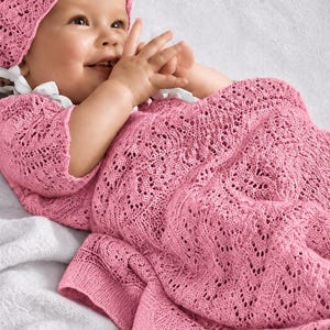 Könnte beinhalten: Ein Set aus einer rosa Strickhaube und einer passenden Decke mit einem komplizierten Lochstrickmuster. Die Haube hat weiße Bänder, die unter dem Kinn gebunden werden, während die Decke ein durchgehendes, strukturiertes Strickmuster aufweist.