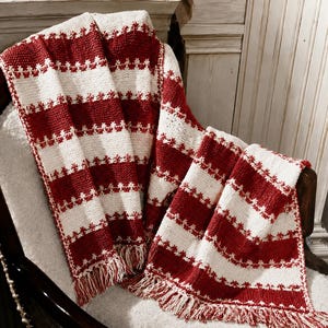 Könnte beinhalten: Eine gestrickte Wohndecke mit abwechselnden horizontalen Streifen aus dunkelrotem und cremeweißem Garn. Das Design enthält ein sich wiederholendes kleines sternartiges Muster zwischen den einfarbigen Bändern und endet mit Fransen am unteren Rand.