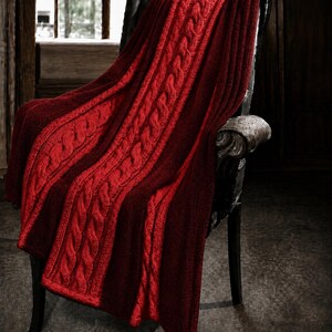 Könnte beinhalten: Eine tiefrote Strickdecke mit Zopfmuster und vertikalen Texturmustern. Die Decke ist über einen dunklen Stuhl drapiert und zeigt eine dicke, gewebte Konstruktion mit abwechselnden Abschnitten aus glattem Strick und erhabenen, gedrehten Zopfmustern.