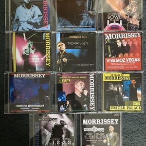 May include: A selection of Morrissey CD albums in clear cases. The covers display images of Morrissey, concert venues, and text with dates and locations. The CDs are arranged in a grid formation.