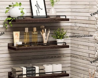 May include: Three wooden shelves with black metal brackets mounted on a wood panel wall. The top shelf has a framed print with black and white calligraphy. The middle shelf has two glass bottles, a small white container, and a set of makeup brushes. The bottom shelf has a stack of books, two rolls of toilet paper, and a white towel.