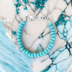 May include: A turquoise beaded bracelet with a silver clasp. The bracelet is on a white and blue marble surface.