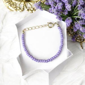 May include: A delicate lavender beaded bracelet with a gold-colored clasp and chain, presented in a white gift box. The bracelet is composed of small, round beads. The background includes purple flowers and a gold decorative element.